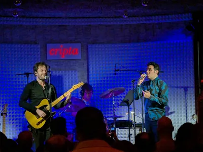 David habla al público con el micrófono; el guitarrista sonríe con los ojos cerrados, todo en luz azul.