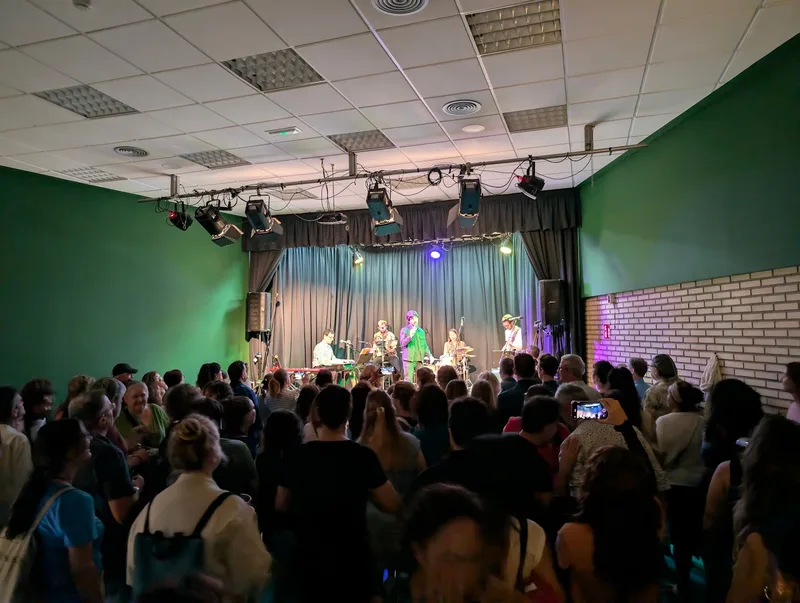 La sala de Loiola Kultur Etxea llena de gente de pie, vista desde el fondo, con el escenario iluminado al final.