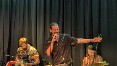 Cantante con coleta y camisa negra de lentejuelas abriendo el brazo hacia el público.
