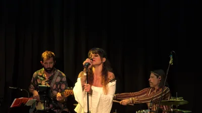 Cantante con blusa blanca de mangas anchas y melena rizada, junto al guitarrista y al batería.