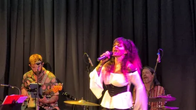 Cantante de blusa blanca y corsé negro con luz rosa, cantando con fuerza.