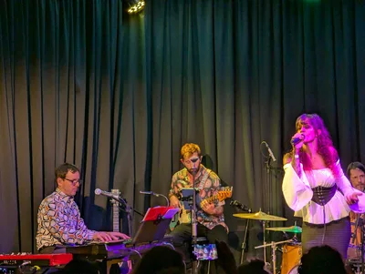 Plano abierto de la banda al completo, con la cantante de blusa blanca en el centro y cabezas del público abajo.