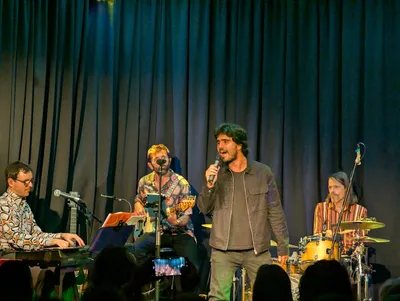 Cantante con cazadora verde y pelo rizado de pie ante el micrófono, con la banda detrás.