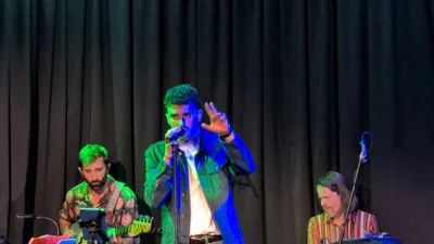 El cantante de cazadora verde bajo luz azul, con una mano levantada.