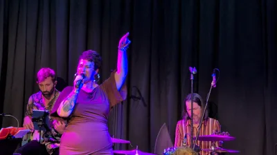 La cantante de vestido malva con el brazo en alto, bajo luz morada.