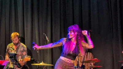Cantante con top negro, brazos tatuados y falda de lentejuelas, señalando con el micrófono.