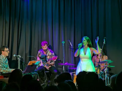 Cantante con vestido blanco largo y una mano en alto, ante la banda y el público.