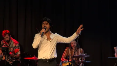 Cantante con camisa blanca abierta y el brazo extendido, a contraluz rojo.