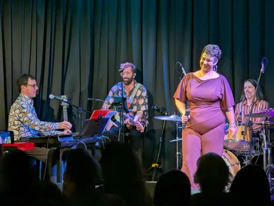 La cantante de vestido malva de pie en el centro del escenario, con el teclista y el batería.