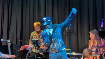 Cantante disfrazado de policía, con gorra de plato, gafas de sol y chaqueta de cuero, con el puño en alto.