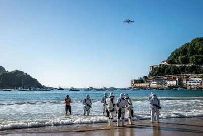 Soldados imperiales con el agua por los tobillos frente al Aquarium y el monte Urgull, con barcos fondeados y un dron encima.
