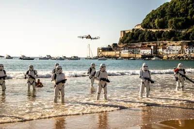 Soldados imperiales avanzan por la orilla entre las olas, uno junto a un bañista; al fondo, Urgull y un dron.