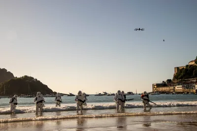 Vista amplia de la bahía al atardecer: una fila de soldados imperiales llega a la orilla y un dron vuela sobre ellos.