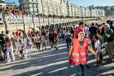 Una voluntaria de Aspanogi, con pañuelo rojo y chaleco, sube por la rampa al paseo de La Concha, lleno de gente en la barandilla.