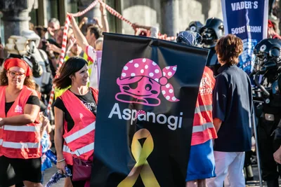 Voluntarias de chaleco rojo junto a la bandera negra de Aspanogi, con su pirata rosa y el lazo dorado del cáncer infantil.