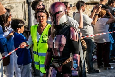 Una guerrera mandaloriana de armadura granate pasa junto al cordón, con el público haciendo fotos con el móvil.