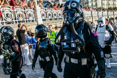 Pilotos imperiales de negro, uno de ellos pequeño, desfilan por el paseo con la barandilla llena de gente detrás.
