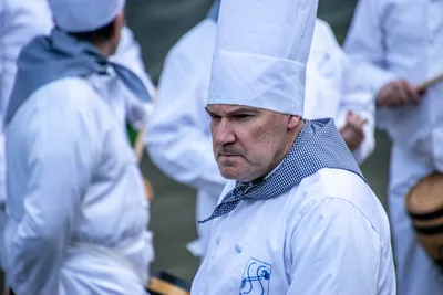 Un cocinero con gorro alto y pañuelo de cuadros azules al cuello, serio entre sus compañeros.