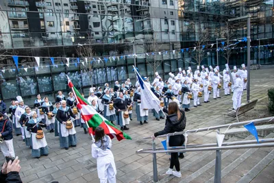 La compañía formada en una plaza de edificios de cristal, con la ikurriña y una bandera blanca y azul delante.