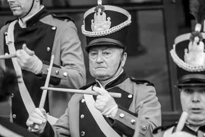 En blanco y negro, un tamborrero veterano con chacó toca con las baquetas en cruz.
