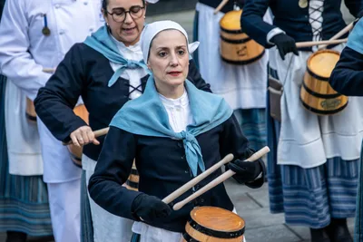 Una aguadora con cofia y toquilla celeste toca la barrica con la mirada al frente.