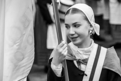 En blanco y negro, una abanderada joven con cofia y banda cruzada sujeta el asta.