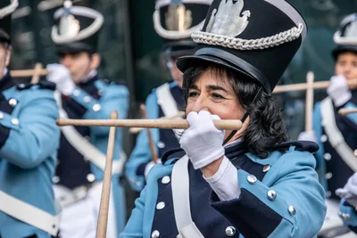 Una tamborrera de azul celeste con guantes blancos cruza las baquetas delante de la boca.