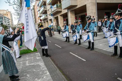 En la calle, una abanderada saluda con la bandera blanca y azul frente al tambor mayor y la fila de soldados.