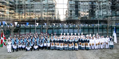 Foto de grupo de toda la compañía delante de un edificio de cristal, con la ikurriña a un lado y la bandera blanca y azul al otro.