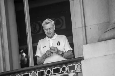 En blanco y negro, un hombre mayor con el polo blanco del club sostiene una copa en una ventana.