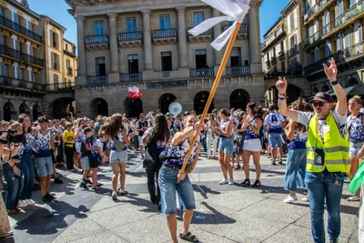 Gente bailando en corro alrededor de la bandera, y un hombre con chaleco amarillo con los brazos arriba.