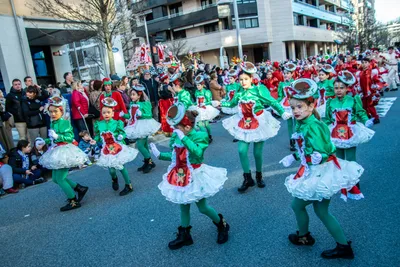 Comparsa infantil de verde y rojo bailando en la calzada, con el público sentado en la acera.