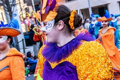 Joven con antifaz blanco, sombrero de plumas y traje naranja y morado de lentejuelas.