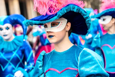 Joven con antifaz blanco y sombrero azul rematado en plumas rosas, con la comparsa azul detrás.