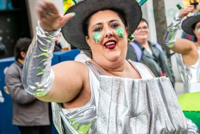 Mujer con sombrero negro de ala ancha y traje plateado, saludando a la cámara.