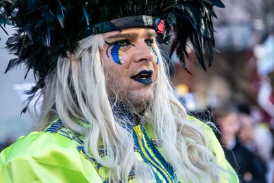 Hombre con peluca blanca larga, tocado de plumas negras y maquillaje azul.
