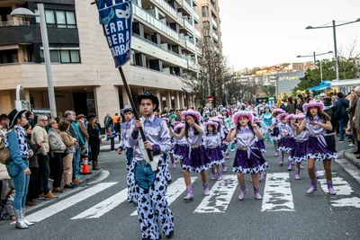 La comparsa del barrio desfila de lila y blanco detrás de su estandarte, con el público a los lados.