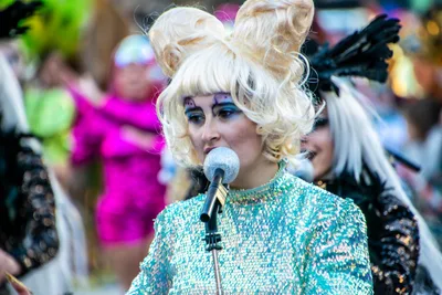 Una cantante con peluca rubia muy alta y traje de lentejuelas canta ante un micrófono.