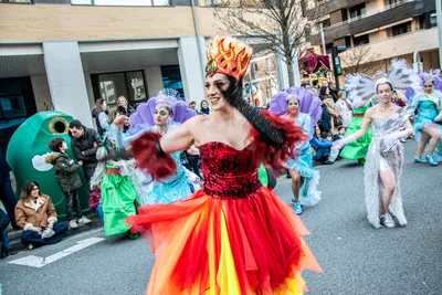 Una bailarina de rojo y naranja avanza por la calzada, con la comparsa de azul y blanco detrás.