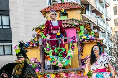 Carroza con una casita de balcón y una figura de abuela asomada, todo lleno de flores de papel.