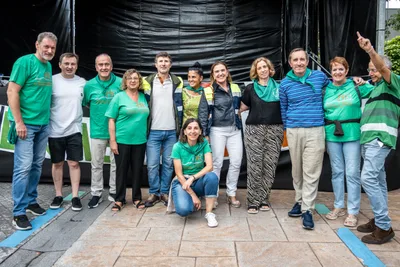Una docena de personas posan abrazadas en el escenario, casi todas con la camiseta verde de las fiestas; una señala hacia arriba.