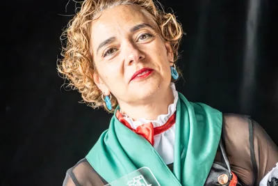 Una pregonera de pelo rizado, con gorguera blanca, lazo rojo y el pañuelo verde al cuello, mira al público.