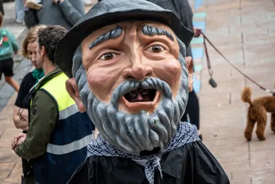 Un cabezudo de anciano con barba gris, boina negra y pañuelo al cuello, muy de cerca, con gente y un perro detrás.