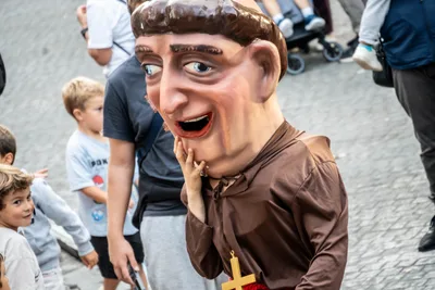 Un cabezudo de fraile con hábito marrón y crucifijo se lleva la mano a la mejilla entre niños que lo miran.