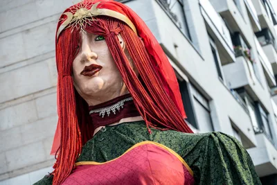 Una giganta pelirroja con diadema dorada y vestido verde y rojo, vista desde abajo entre los edificios.