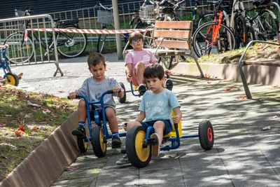 Tres niños pequeños pedalean en karts de plástico por un camino de la plaza.