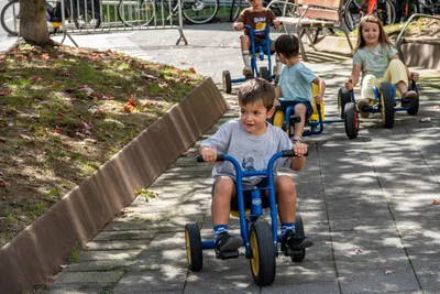 Un niño pequeño en un triciclo azul, con otros dos detrás, a la sombra de los árboles.