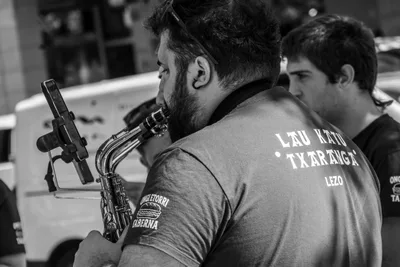 En blanco y negro, un saxofonista de espaldas con «Lau Katu Txaranga · Lezo» en la camiseta.