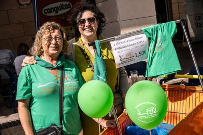 Dos mujeres sonríen en el puesto de pañuelos y camisetas de las fiestas, con globos verdes y el cartel del pañuelo a 1,50 euros.