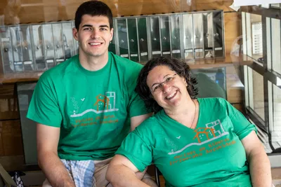 Un chico y una mujer con la camiseta verde de la asociación sonríen sentados junto a los buzones de un portal.